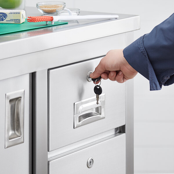 A hand using a key to unlock a drawer on a Regency stainless steel table with drawers.