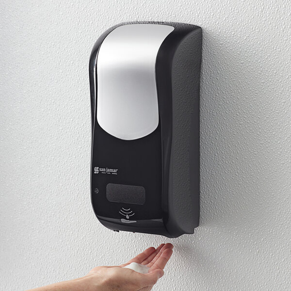 A hand using a black and silver San Jamar Summit Rely hybrid automatic foam soap and sanitizer dispenser.