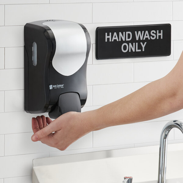A black and silver San Jamar manual hand soap, sanitizer, and lotion dispenser on a white wall.