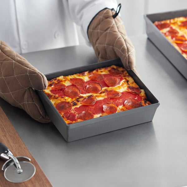 A person holding a Detroit-style pizza in an American Metalcraft hard coat anodized aluminum pan.