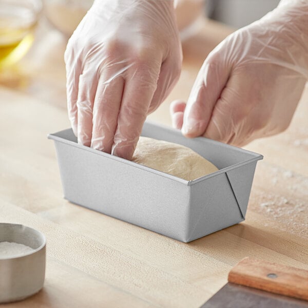 A person wearing plastic gloves kneads dough in a Baker's Mark aluminized steel bread loaf pan.