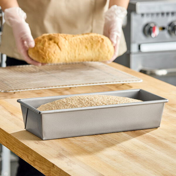 A rectangular glazed aluminized steel bread loaf pan with a partially risen loaf inside, placed on a wooden countertop.