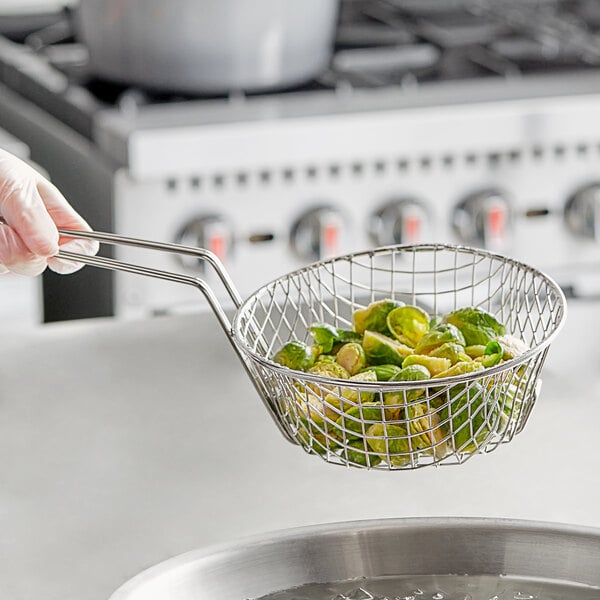 A person holding a Choice round wire culinary basket full of brussels sprouts over a pan.