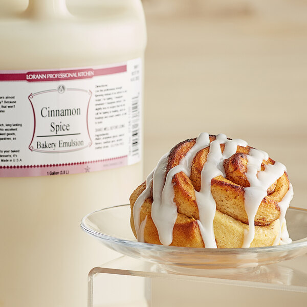 A cinnamon roll on a plate next to a white bottle of LorAnn Oils Cinnamon Spice Bakery Emulsion.