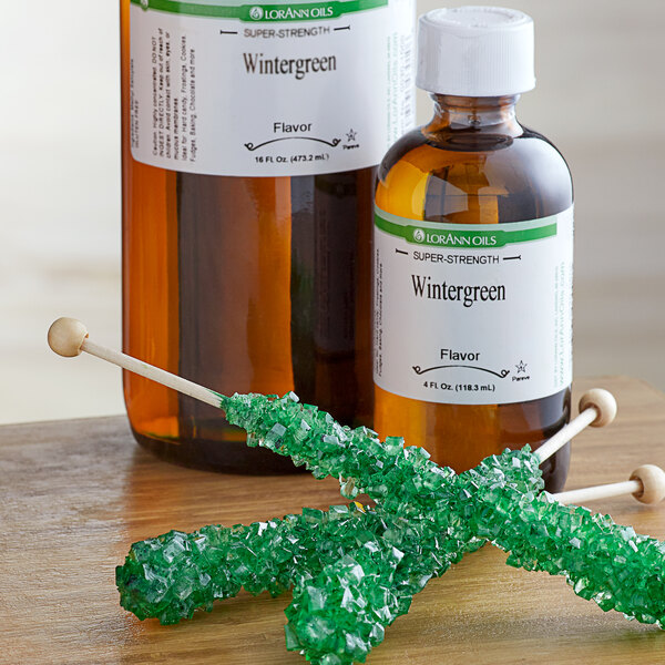 A bottle of LorAnn Oils Wintergreen Super Strength Flavoring on a wooden surface next to green sugar.