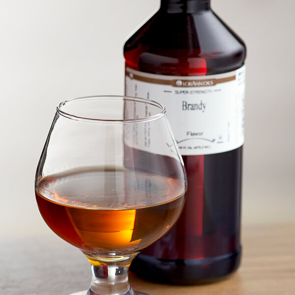A bottle of LorAnn Oils Brandy Super Strength Flavor next to a glass of brown liquid.