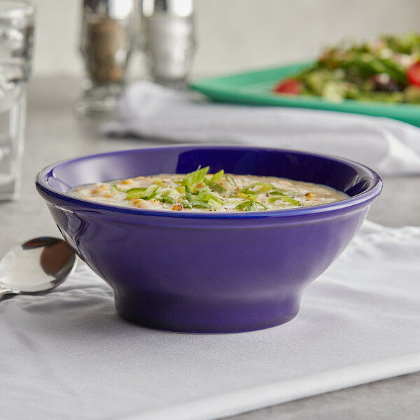 A Tuxton cobalt blue bowl of soup with a spoon on a table with a plate of salad.