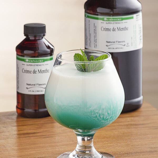 A glass of blue drink with a green leaf on top next to a bottle of LorAnn Oils All-Natural Creme de Menthe Flavor.