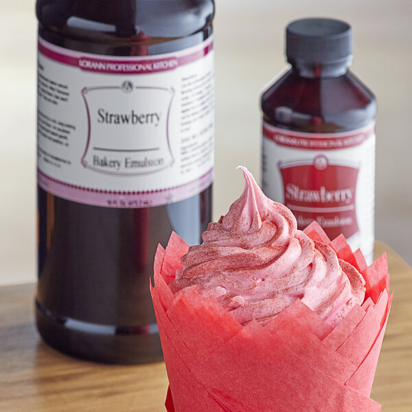 A cupcake with pink strawberry frosting next to a bottle of LorAnn Strawberry Bakery Emulsion.