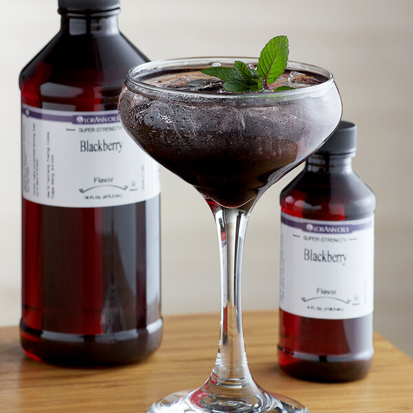 A bottle of LorAnn Oils Blackberry Super Strength Flavor on a table with a glass of black liquid and a leafy green garnish.