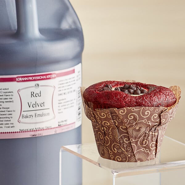 A cupcake in a brown wrapper with red frosting and chocolate chips next to a jug of LorAnn Oils red velvet bakery emulsion.