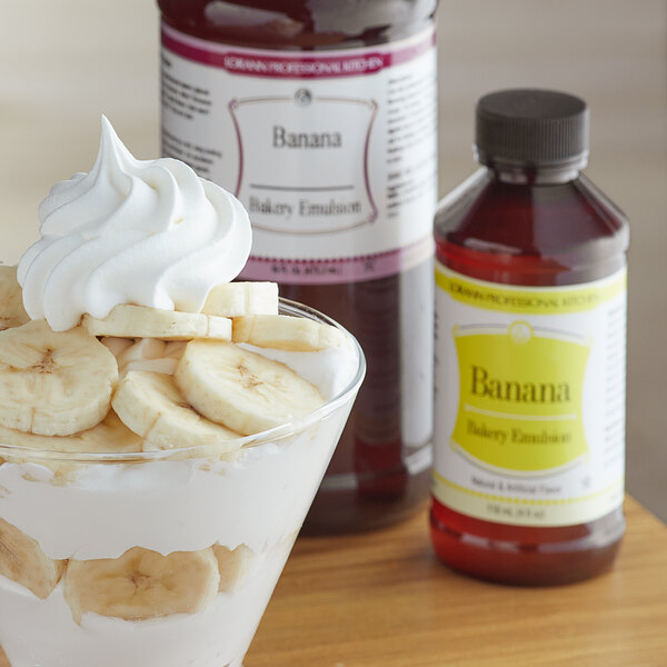 A close-up of a bottle of LorAnn Oils Banana Bakery Emulsion with a banana and banana pudding in the background.