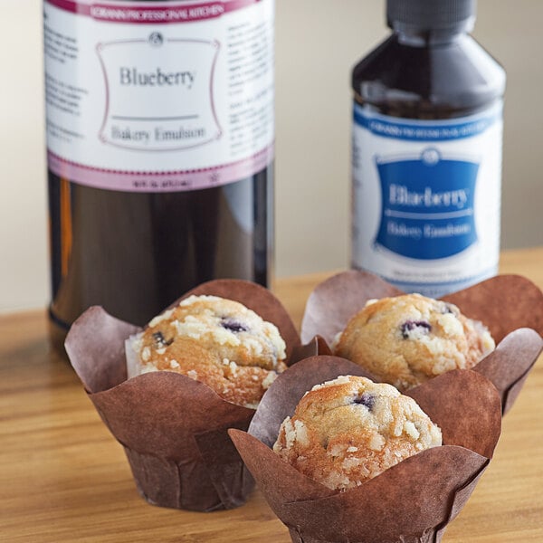 A group of blueberry muffins in brown wrappers next to a bottle of LorAnn Oils Blueberry Bakery Emulsion.