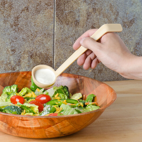 A hand using a Cambro beige plastic ladle to pour dressing over a bowl of salad.