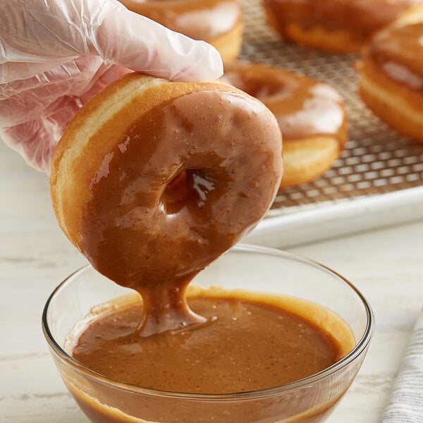 A hand holding a glazed doughnut over a bowl of brown Rich's Maple Heat 'n Ice.
