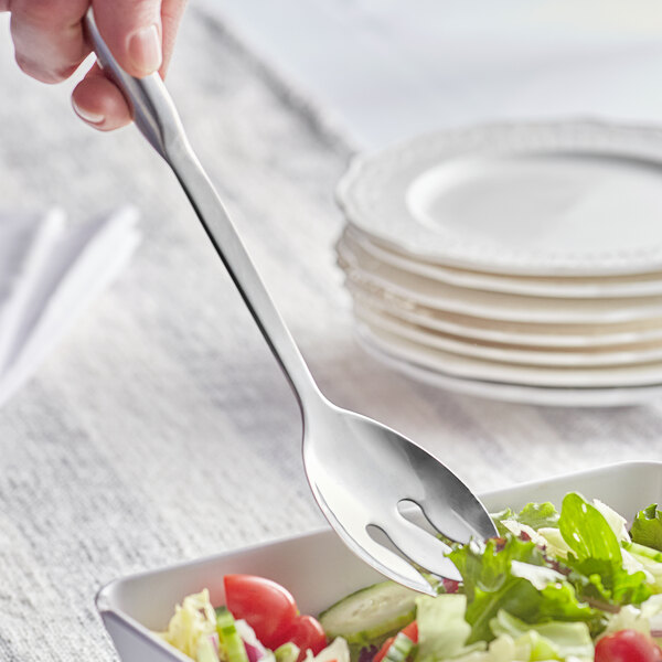 A hand holding a Choice stainless steel notched salad serving spoon over a bowl of salad.