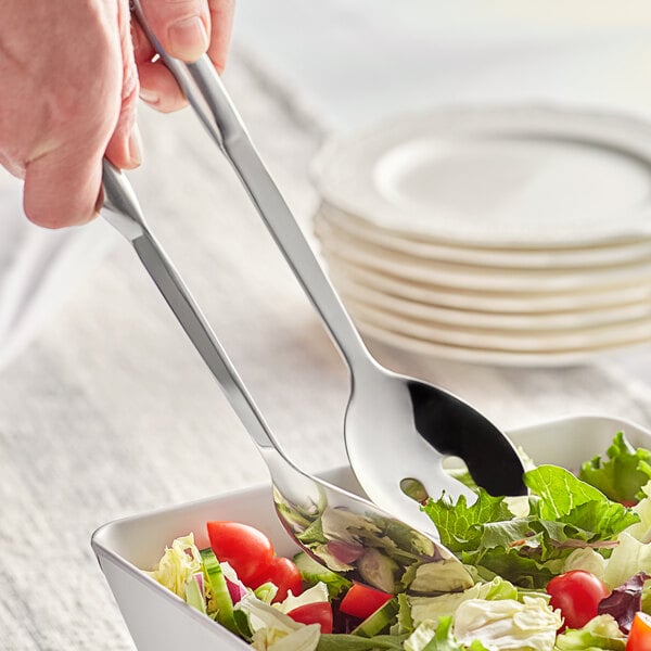 A hand holding a Choice stainless steel salad serving utensils set over a bowl of salad.