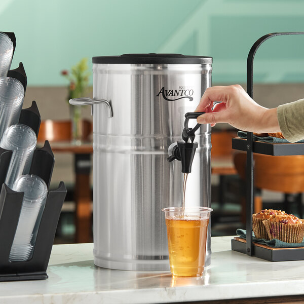 A 2-gallon round stainless steel iced tea dispenser with a spigot, shown dispensing tea into a plastic cup.