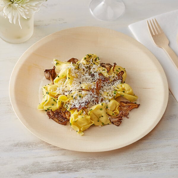 A TreeVive by EcoChoice wooden round plate with pasta, mushrooms, and cheese with a wooden spoon on a white table.