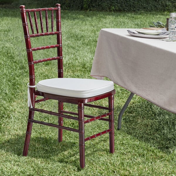 A Lancaster Table & Seating mahogany wood Chiavari chair with an ivory cushion next to a table with a white tablecloth.