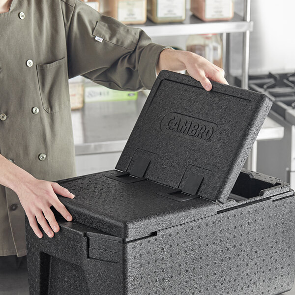 A man in a chef's jacket opening a black Cambro box lid on a counter in a professional kitchen.