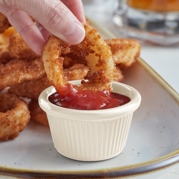 A hand dipping a fried onion ring into a small white Acopa fluted ramekin of sauce.