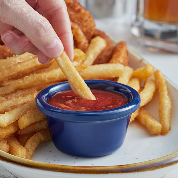 A hand holding a french fry being dipped into a blue Acopa ramekin of ketchup.