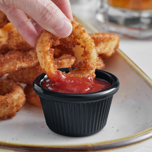 A hand dipping a fried onion ring into a black Acopa ramekin of red sauce.