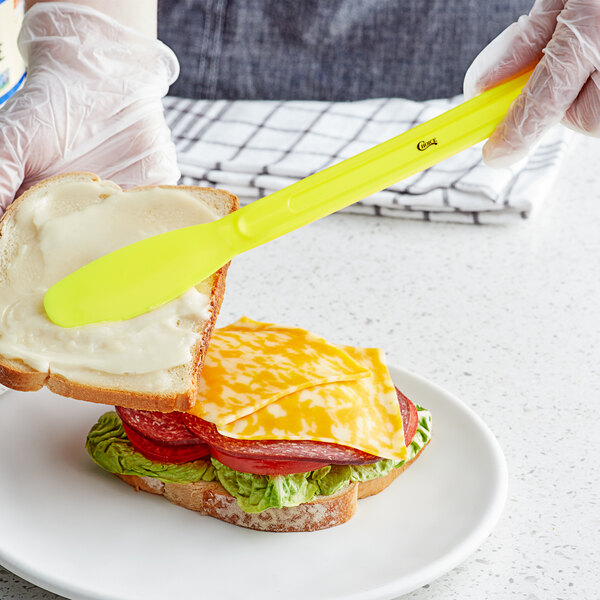 A person using a Choice neon yellow sandwich spreader to spread butter on a sandwich.