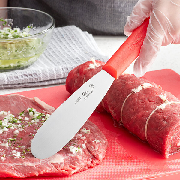 A person using a Choice stainless steel sandwich spreader to cut meat on a red cutting board.