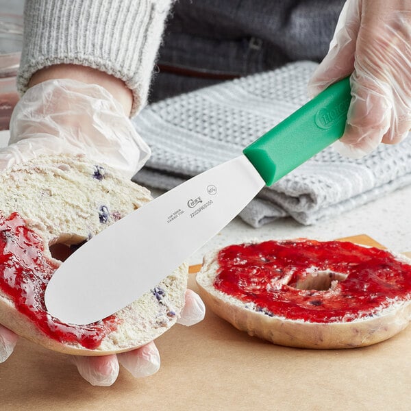 A person spreading jam on a bagel with a Choice green stainless steel sandwich spreader.