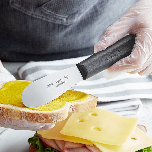 A person using a Choice stainless steel sandwich spreader with a black handle to spread butter on a piece of bread.