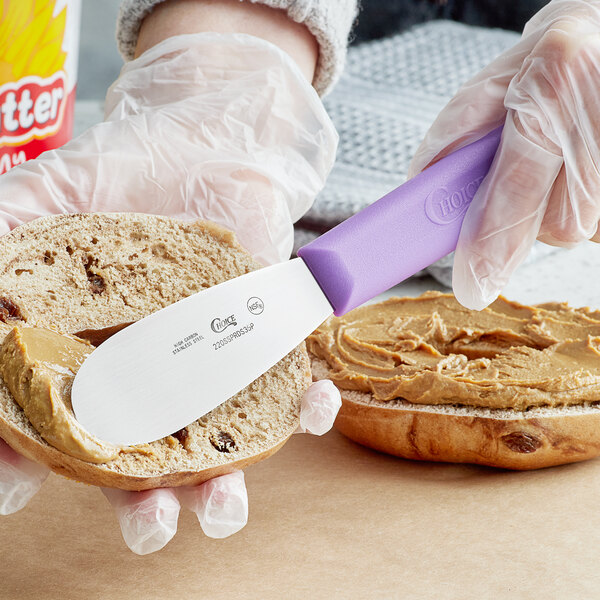 A person spreading a sandwich spread with a Choice stainless steel sandwich spreader with a purple handle on a piece of bread.