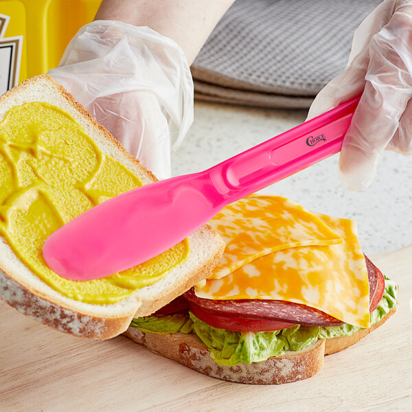 A gloved hand uses a Choice neon pink polypropylene sandwich spreader to spread butter on a piece of bread.