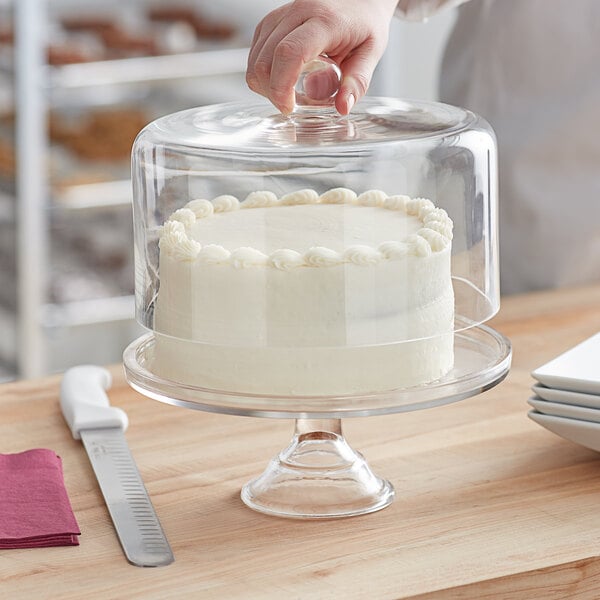 A person using an Acopa clear glass cake stand with a cover to serve a white frosted cake.