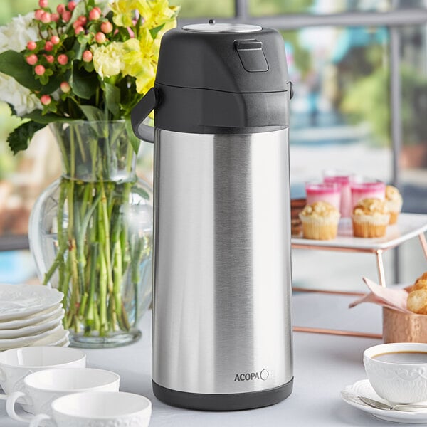 An Acopa stainless steel coffee airpot on a table with a white cup filled with coffee.