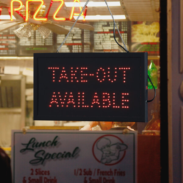 A rectangular LED sign that says "Take Out Available" on a counter in a pizza parlor.