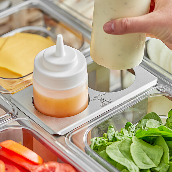 A person pouring dressing from a Server stainless steel squeeze bottle holder into a container on a salad bar.