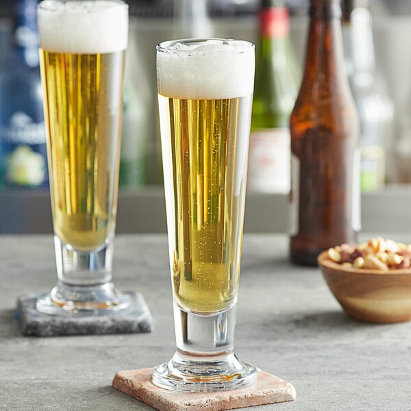 Two Pasabahce Cin Cin footed pilsner glasses of beer on a table with a bowl of nuts.