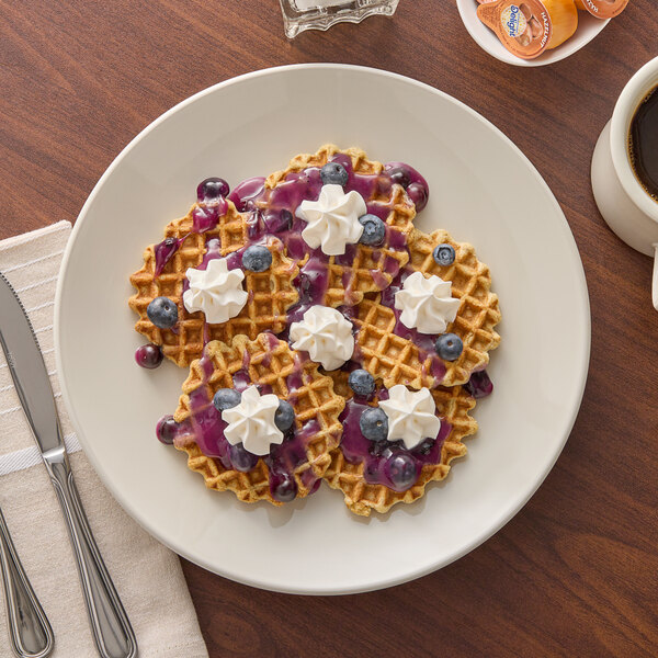 An Acopa ivory stoneware plate with waffles, whipped cream, and blueberries on a table.