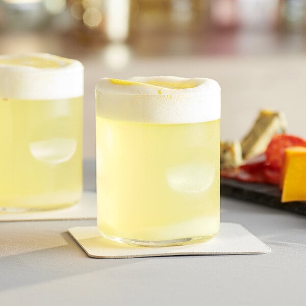 Two Acopa thumbprint juice glasses filled with yellow liquid and foam.