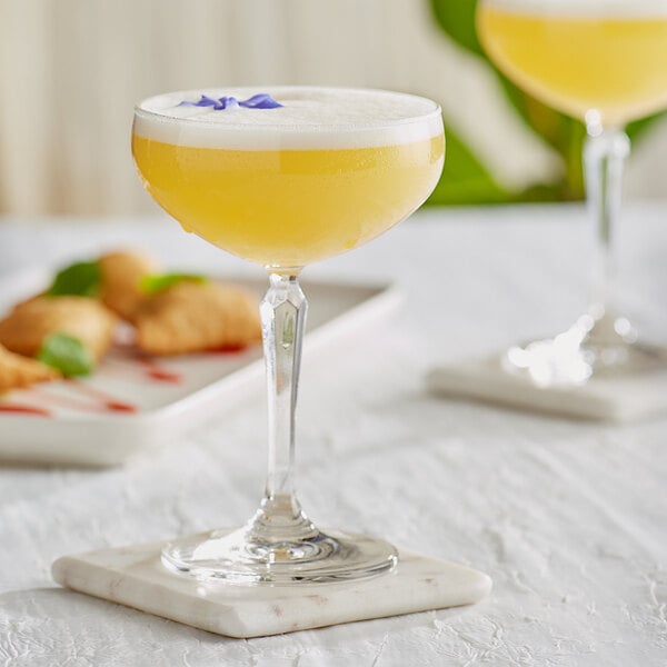 A coupe glass of yellow liquid on a white marble stand with a garnish.