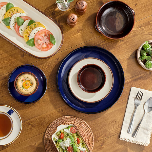 An 8 oz. vanilla bean stoneware mini casserole dish with lid is displayed on a wooden table among various plates and food items.