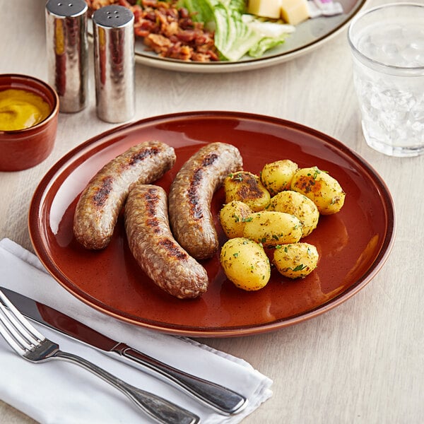 A plate of sausages and potatoes on an Acopa Sedona Orange stoneware coupe plate with a knife and fork.