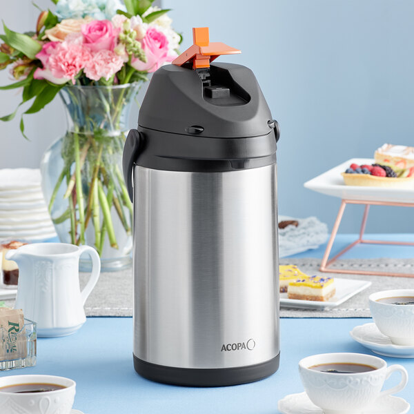 An Acopa stainless steel coffee airpot with a metal lever on a table with a cup of coffee.