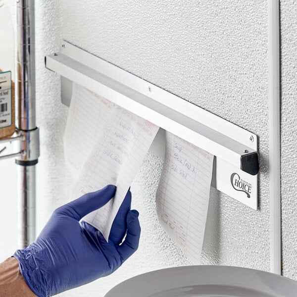 A person wearing blue gloves putting a piece of paper in a Choice aluminum wall mounted ticket holder.