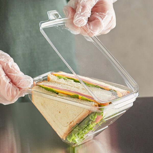 A gloved hand holding a clear PET sandwich container with a sandwich inside.