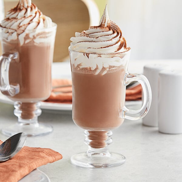 Two Acopa glass Irish coffee mugs filled with hot chocolate and whipped cream on a table with a spoon and napkin.