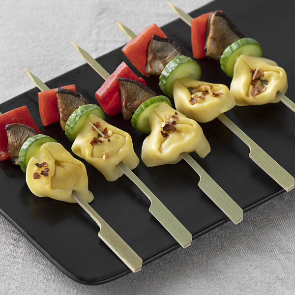 Bamboo skewers of tortellini, bacon, and green peppers on a black plate.