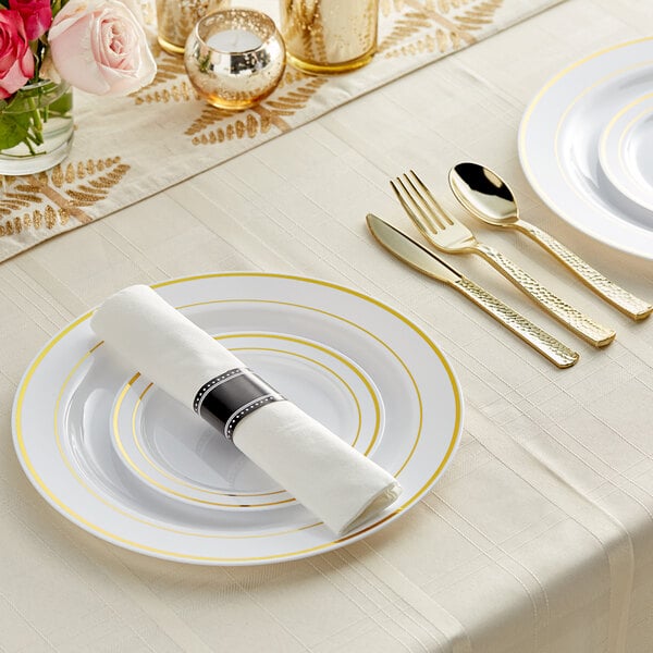 A white table set with Visions gold banded plastic dinnerware and rolled hammered flatware.